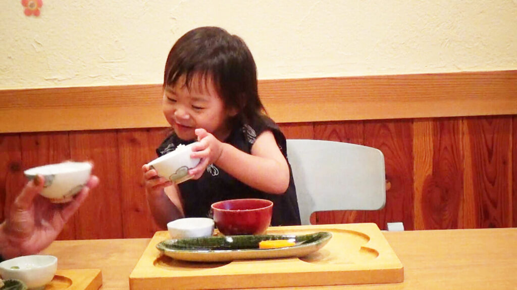 幼児が食事の茶碗を保護者に楽しそうに見せている様子