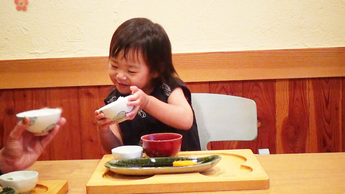 幼児が食事の茶碗を保護者に楽しそうに見せている様子